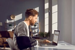 Man with broken arm working on laptop computer in office. Side view shot of businessman wearing arm splint sitting at desk in front of window working distantly at home