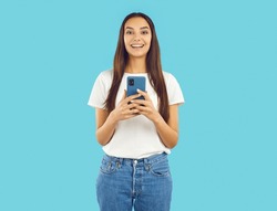 Joyful excited young woman using mobile phone for online shopping, chatting, watching media or playing games. Girl with smiling shocked expression holding smartphone isolated on light blue background.