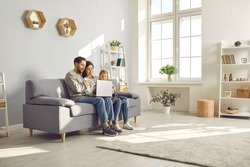 Happy family spend time in spacious room at home. Couple with child watch online movie on laptop sitting on sofa in cozy light living place space with white grey walls, shelves, house plants and wifi