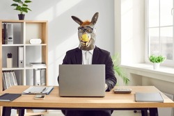 Funny business man and company employee wearing suit and animal donkey mask sitting at the desk on his workplace and working on a laptop in office. Entertainment, fun and fools day concept.