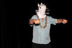 An unidentifiable man wears a White Rubber Horse Head Mask, and a Gold Chain while in a Photo Booth with black velvet curtains as a background. With room for your text. Photo Booths are popular fun. 