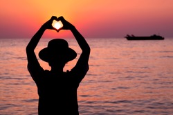 silhouette Woman sends a heart-shaped hand signal on sunset in sea background.