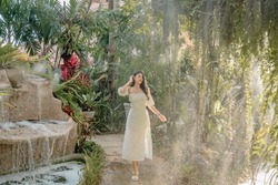 Young woman walking in tropical garden in long summer dress, greenery and trees around, enjoying nature