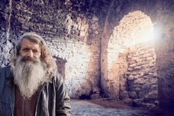 In the subterranean depths of a castle, an old man with a silver beard stands against a stone wall, illuminated by sunlight streaming through a small window, creating an enigmatic atmosphere