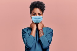 Gorgeous dark skinned young female with Afro hairstyle wearing protective mask against COVID virus, tired of stress and tension, looks confidently at the camera, poses against beige studio background