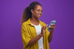 Young optimistic long haired African American woman using mobile phone with slight smile dialing friend number wants to make call to gossip or arrange date stands on lilac studio background