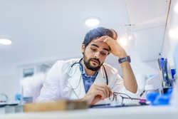 Overworked doctor in his office. Tired male scientist trying to focus, rubbing his forehead with fingers. Mid adult male doctor working long hours. Stressed male doctor sat at his desk 