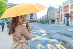A girl with a yellow umbrella is hiding from the rain. Beautiful woman with umbrella on a rainy day. Attractive young woman carrying umbrella and smiling while standing on the street