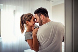 Handsome man in white T-shirt carrying young attractive woman while spending free time at home. Loving young hipster couple relaxing at home and kissing. View from another room through the doorway