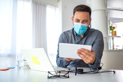 Man in a medical mask at the office. Man works remotely. The guy uses a tablet PC and a laptop for work, and medicine mask and antiseptic for self protective. Coronavirus pandemic, influenza, covid19