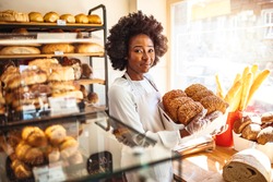 Happy business owner at a bakery shop. Young bakery owner holding a tray with bread in her shop. Beautiful baker. Happy woman working at the bakery and looking at the camera smiling - small business