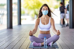 Woman with a protective mask does meditation after training sitting on a fitness mat. Meditating woman wearing a medical face mask to protect from corona virus covid-19 with eyes closed on a yoga mat.