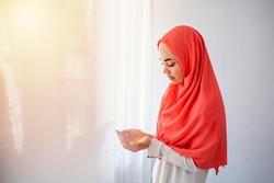 Muslim woman in hijab praying indoors. Muslim woman in beige hijab and traditional clothes praying for Allah, copy space. Muslim woman with hijab praying indoor at bright window. 