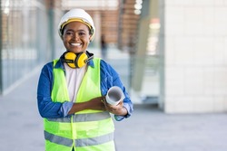 Portrait of woman engineer at building site looking at camera with copy space. Mature construction manager standing in yellow safety vest and white hardhat with crossed arms. 