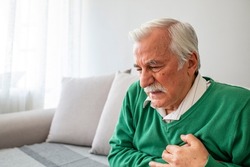 Senior man suffering from heart attack at home. Closeup shot of a mature man holding his chest in discomfort oindoors. Worried senior man feeling unwell and having chest pain in the living room.