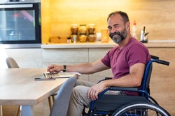 Happy Mature Man in wheelchair from home. Cheerful businessman smile while sitting in his wheelchair. Disabled Man in Wheelchair using Laptop At Home. Mature man sitting on wheelchair at the table.