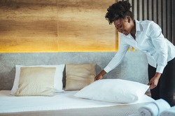 Maid working at a hotel making the bed and pillows. Maid making bed in hotel room. Housekeeper Making Bed. Young maid making bed in light hotel room.