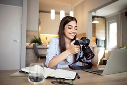 Happy young photographer holding a dslr camera in her home office. Female photographer smiling cheerfully while working at her desk. Creative female freelancer working on a new project.