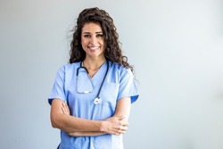 Modern Medical Education Concept. Portrait Of Smiling Female Doctor In Blue Coat Posing With Folded Arms Over Light Background. Portrait Of Female Nurse Wearing Scrubs In Hospital