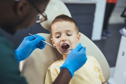 Dentist doing regular dental check-up to little boy. Little cute smiling boy is sitting in dental chair in clinic, office. Doctor is preparing for examination of child teeth with tools. 