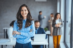 Successful businesswoman standing in creative office and looking at camera. Young woman entrepreneur in a coworking space smiling. Portrait of beautiful business woman standing in front of team