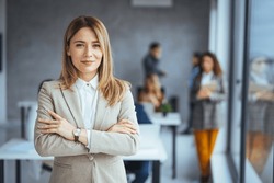 Successful businesswoman standing in creative office and looking at camera. Woman entrepreneur in a coworking space smiling. Portrait of beautiful business woman standing in front of business team