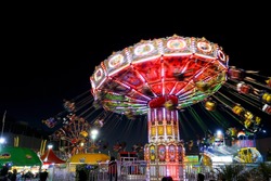 Carnival amusement part with rides that are lit up with colored lights. Blurred fun bright and colorful background. Summer fair at night. Spinning swings with people.