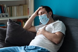A sick caucasian Man wearing face mask feeling sick headache and cough because of Coronavirus Covid-19 on the sofa in quarantine