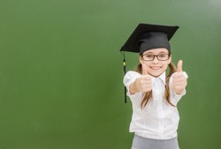 Happy girl in graduation cap near a school board showing thumbs up. Space for text