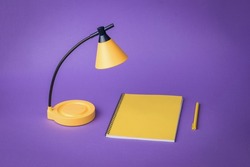 Minimal concept. Yellow desk lamp, yellow notebook and pen on a purple background. Desktop lighting.