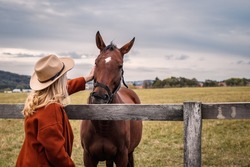 Woman wearing cowboy hat stroking her horse at ranch. Cowgirl at pasture in animal farm. Friendship between people and horse