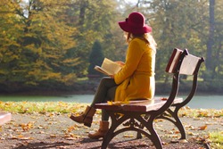 Woman reading a book on a park bench, leisure time in autumn park. fashion woman in yellow coat sitting on a bench. 