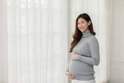 Happy Pregnant Woman turtleneck sweatshirt standing in front of windows and stroking big belly with love at cozy home,Pregnancy of young woman enjoy with future life,Motherhood and Pregnant Concept