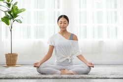 Calm of wellness Asian young woman sit on carpet breathing with yoga lotus pose,Yoga meditation of young healthy woman relax and comfortable at white cozy home,Yoga Exercise for Wellness Concept