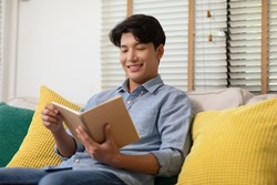 Happy handsome asian man sit on couch smile and reading book relax time at home.Cheerful businessman dressed in blue shirt sitting on sofa and reading a book