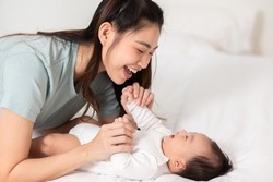 New asian mom playing to adorable newborn baby on bed smiling and happiness at home.Mom talking with infant baby and holding her hands laughing together.Baby and Mother day concept