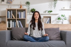 Clam of Asian young woman doing yoga lotus pose to meditation and relax on couch during work online at home.Happiness female break after worked close her eyes and deep breath with yoga so peaceful.
