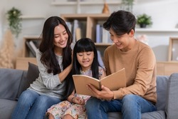 Happy Asian family sitting on the sofa together looking at daughter's note book. Parents are smiling, reading daughter book and proud of education progress. Watching book together, family relationship