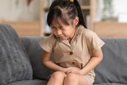 little Asian girl have a stomach ache on couch at home. Child squeeze on her belly so pain and illness. kid has a grimace face. Child health care