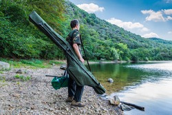 Fishing adventures, carp fishing. Fisherman on a lake shore with camouflage fishing gear, green bag and rod holdall