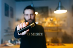 Young beard man show vaping device on his outstretched hand in camera. Selective focus. Vaping concept. Copy space.
