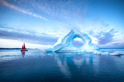 Sail boat with red sails cruising among massive ice bergs during dusk. Disko Bay, Greenland.