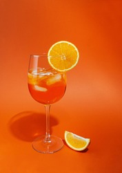 Cocktail aperol spritz with ice. In a glass goblet. On a glass of orange slice. Next to the glass is a slice of orange. On a plain orange background. Hard light, sharp shadows. Close-up Place for tex