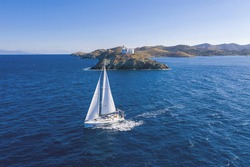 Sailing. Sailboat with white sails, rippled sea background, Lighthouse on a cape. Greece, Kea Tzia island. Summer holidays in Aegean sea. Aerial drone view