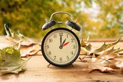 Fall Back one hour. Daylight Saving Time, Black alarm clock with time change on wooden table. Autumn trees and leaves background