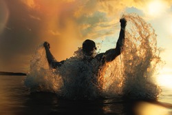 Strong and athletic man jumps out of the water at sunset, flying a lot of splashing