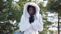 sad dark-skinned woman trembles with cold and warms hands against green pine-trees in cold winter close-up