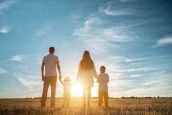 Family with children sons stands joining hands along shadowed field at back setting sun in summer under blue sky backside view