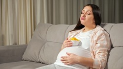Pregnant woman with long brunette hair enjoys eating junk unhealthy fast food from bowl put on large belly sitting on sofa in living room.