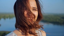 Wind plays with flying locks of pretty brown haired young woman with light makeup posing for camera on tranquil river bank on summer day closeup
